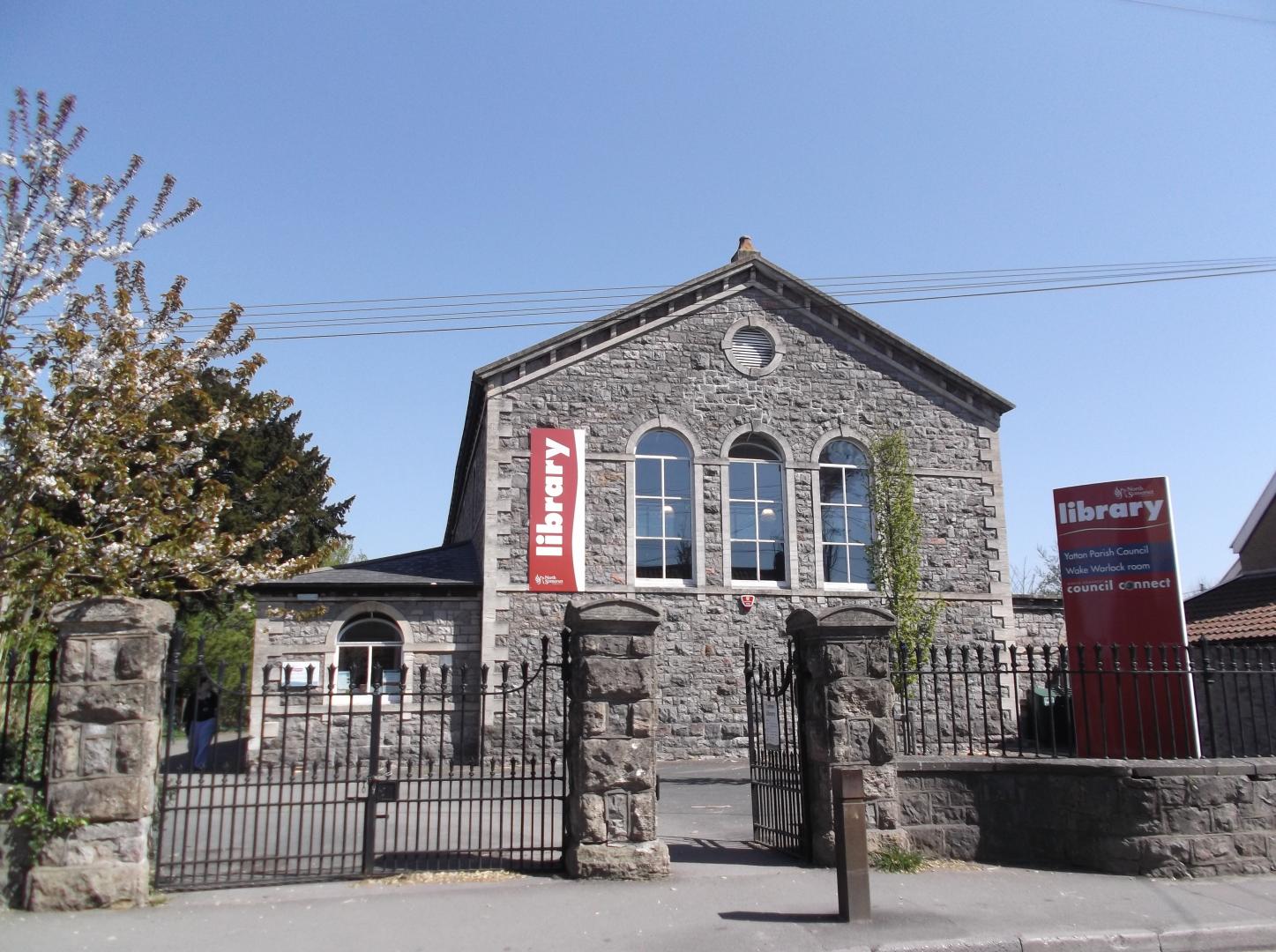A picture of a grey stone building with three windows, an iron fence and a tall red sign