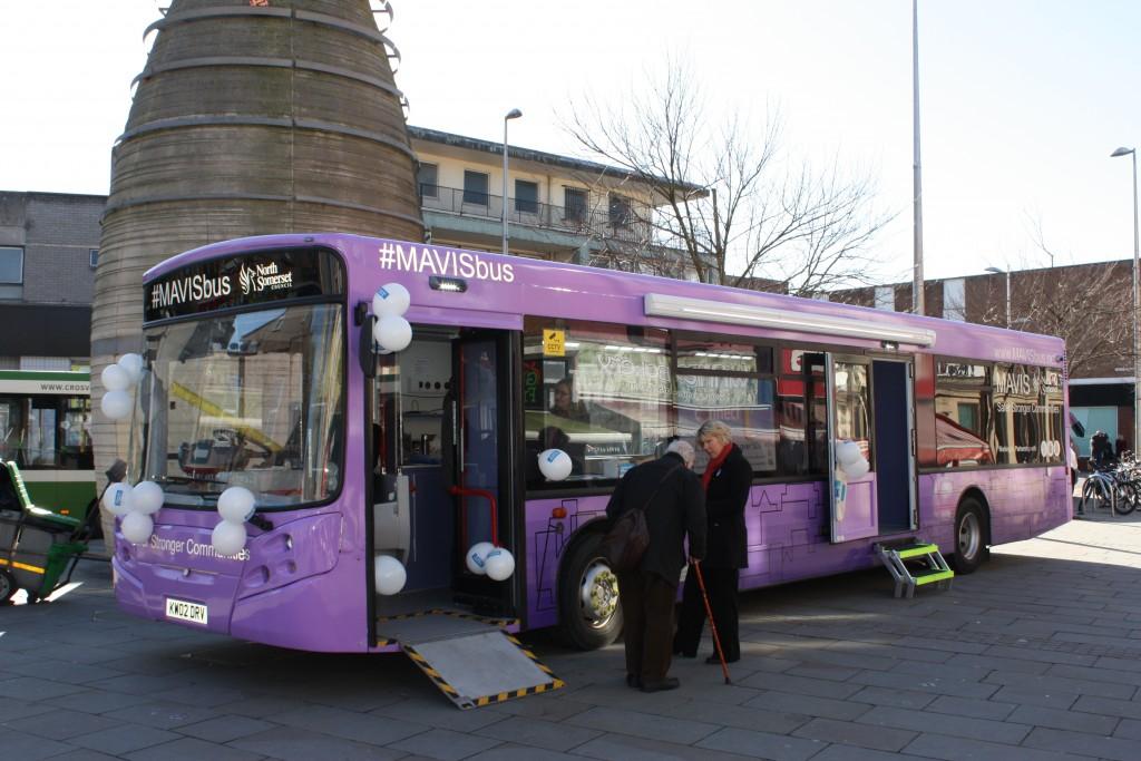 A woman helps an elderly woman onto a purple bus in the town centre