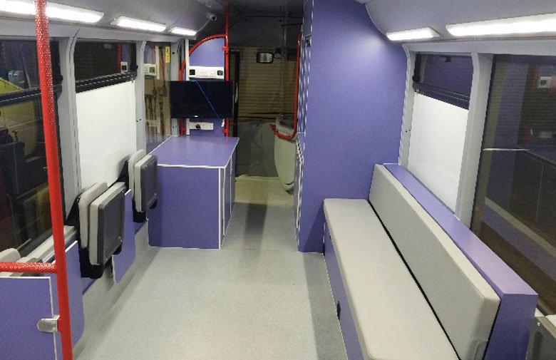An inside view of MAVIS bus, with a grey seated bench and seats and purple room dividers. 