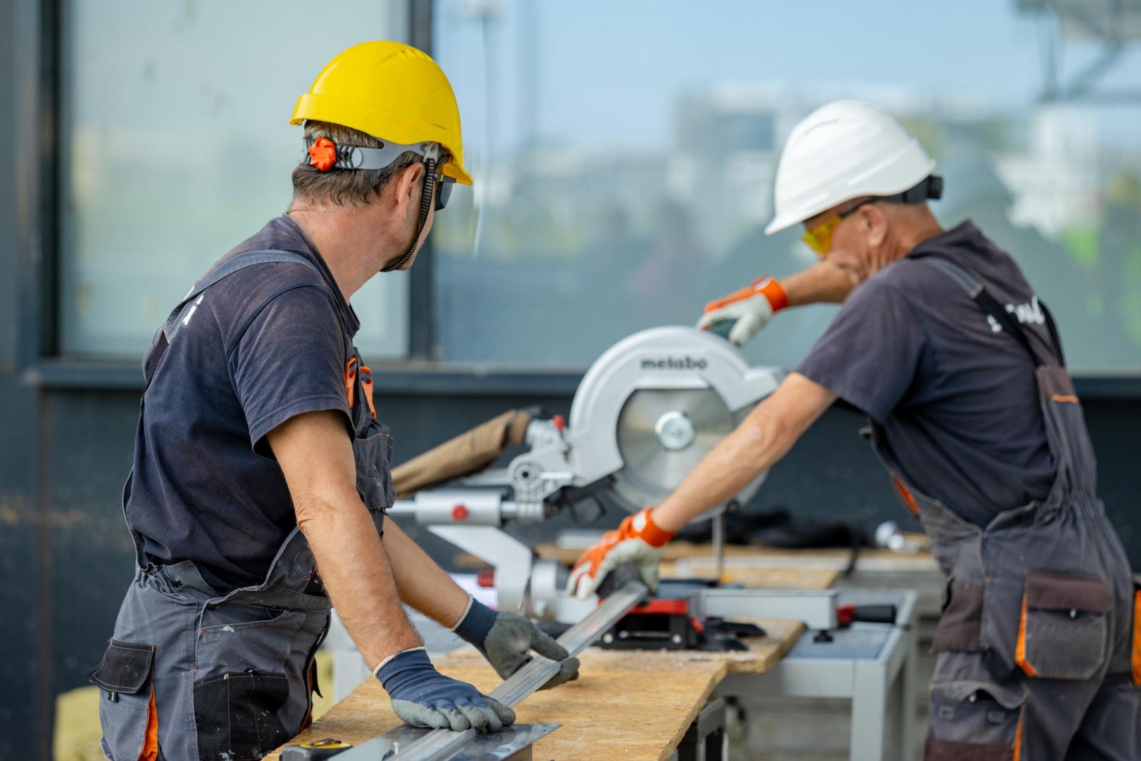 Two workers in hard hats cutting a metal beam with a circular saw