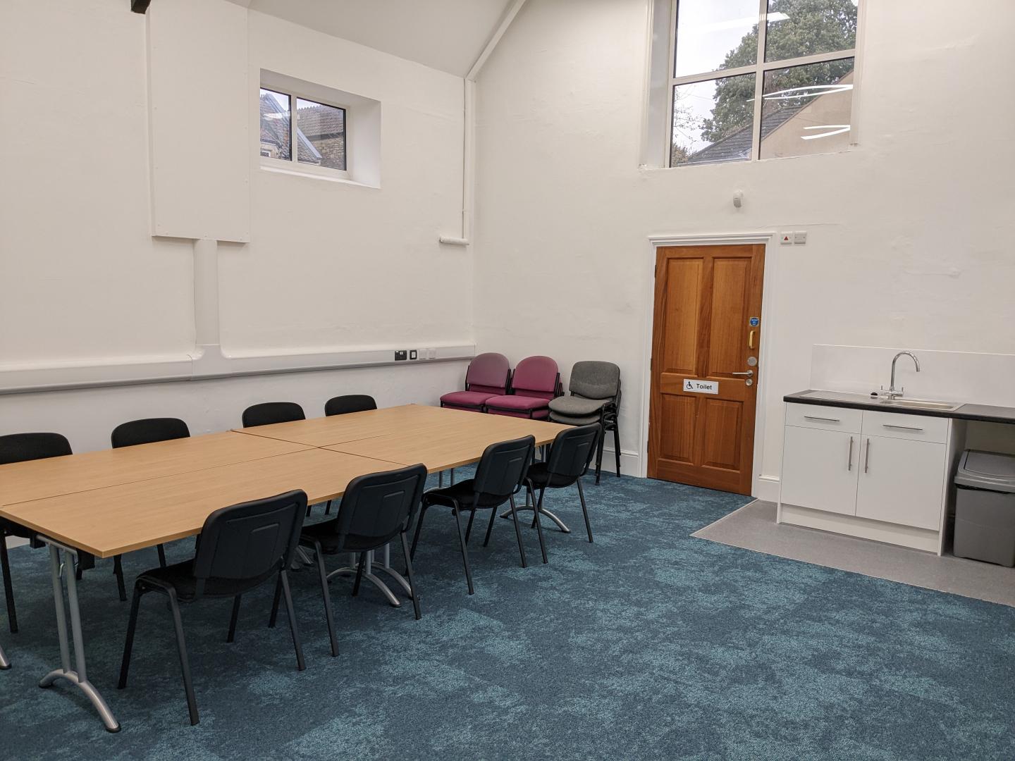 An interior of a meeting room with a table in the centre surrounded by black chairs. There is a sink in the corner of the room and a door to a disabled toilet.