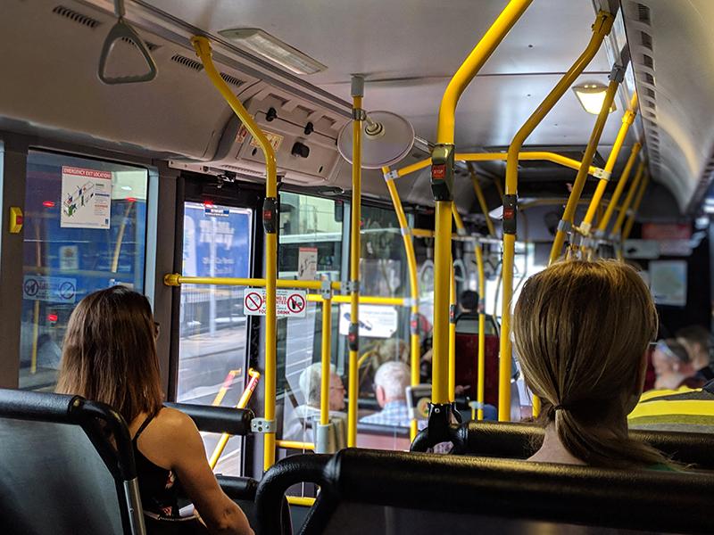 An image of people inside of a bus, seated and standing while holding onto the yellow support bars