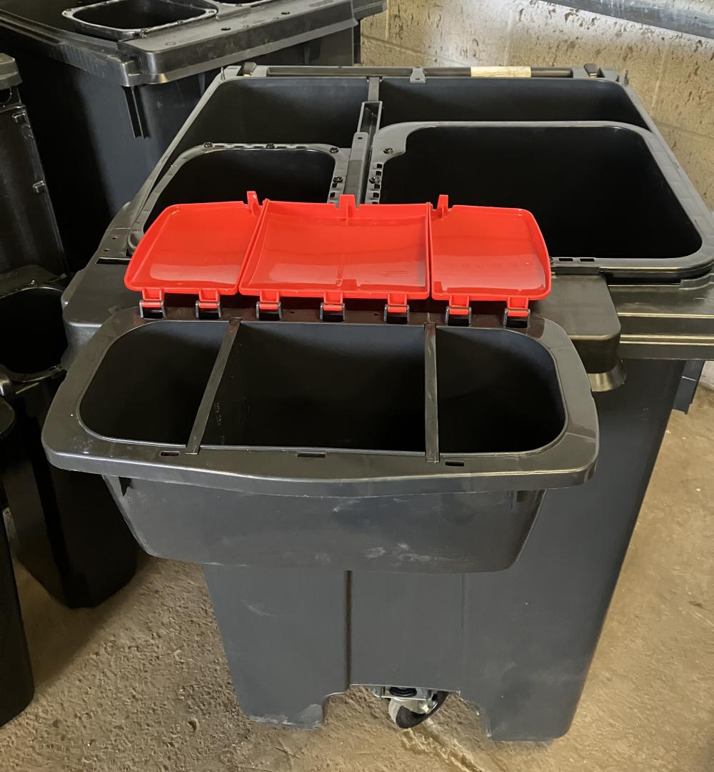 A black wheelie bin divided into four compartments. It also has a fifth compartment which is hooked on the front with a red lid.