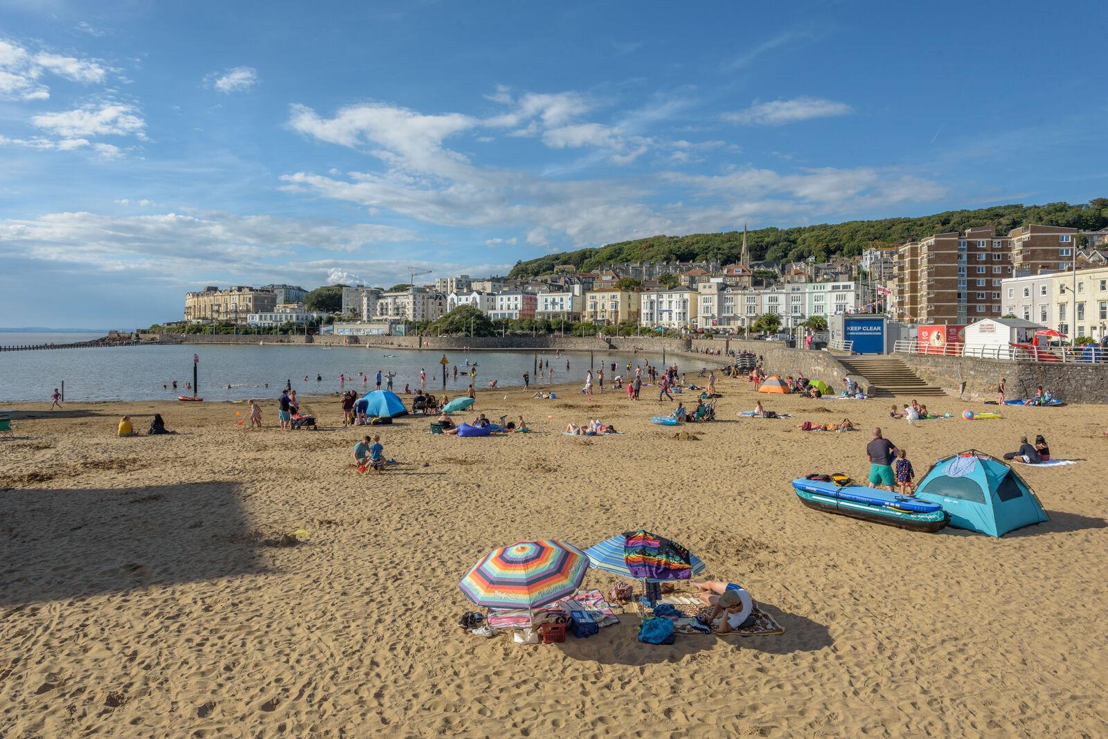 A wide angle shot of Marine Lake in Weston-super-Mare. A wide stretch of beach under a blue sky with a small lake visible to the left. People are dotted about on towels and swimming in the water. 