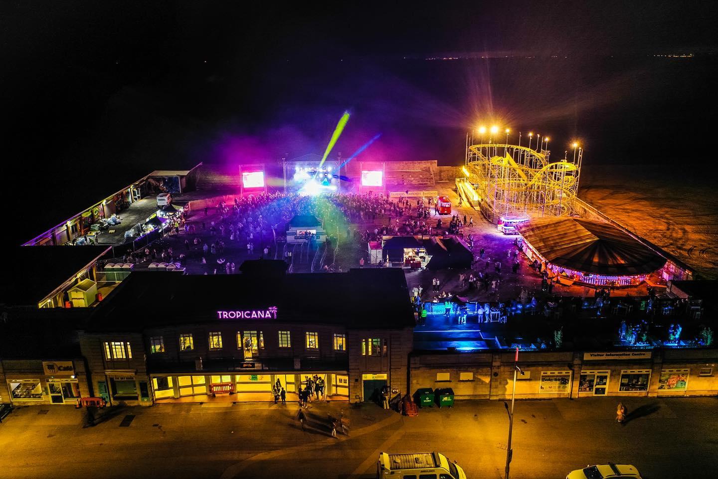 A overhead shot at night of the Tropicana as it was when Banky's Dismaland was here in 2015. There are multi-coloured bright lights everywhere, and people lined up outside the front of the venue.