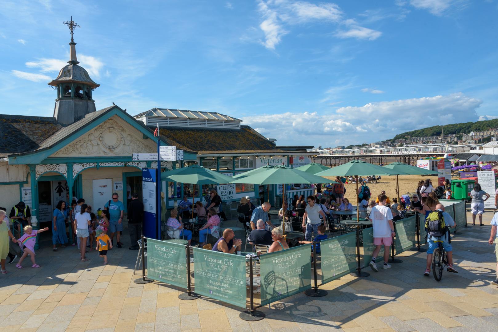 The Victorian cafe on a sunny day. People sit outside the building under umbrellas, and the beach is visible in the background
