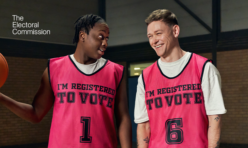 Two young male basketball players are wearing red practice vests which say “I’m Registered to Vote” on them. One player is looking at the other, who is smiling.