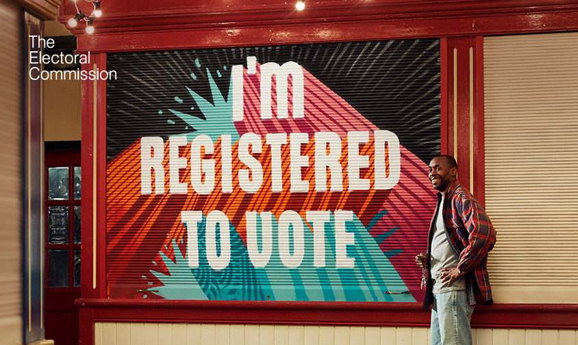 A shopkeeper is standing outside his market shop. The shutters have been pulled down, and “I’m Registered to Vote” has been painted on them in a street art style.