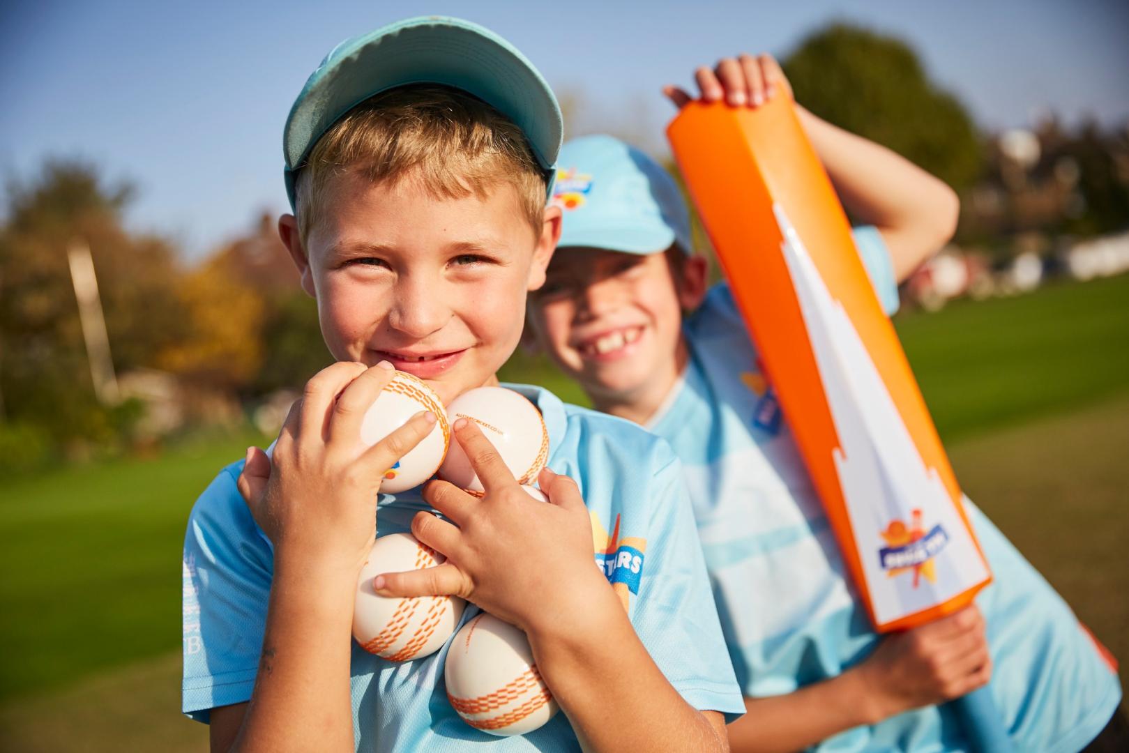 A boy holds cricket balls in his arms and smiles at the camera, while another boy stands behind him holding a cricket bat