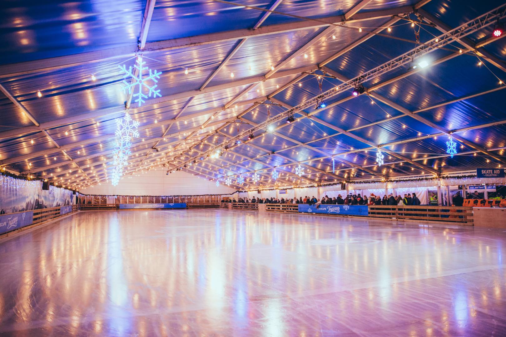 A large covered ice rink, with festive lights on the ceiling and skaters lining the wall. 