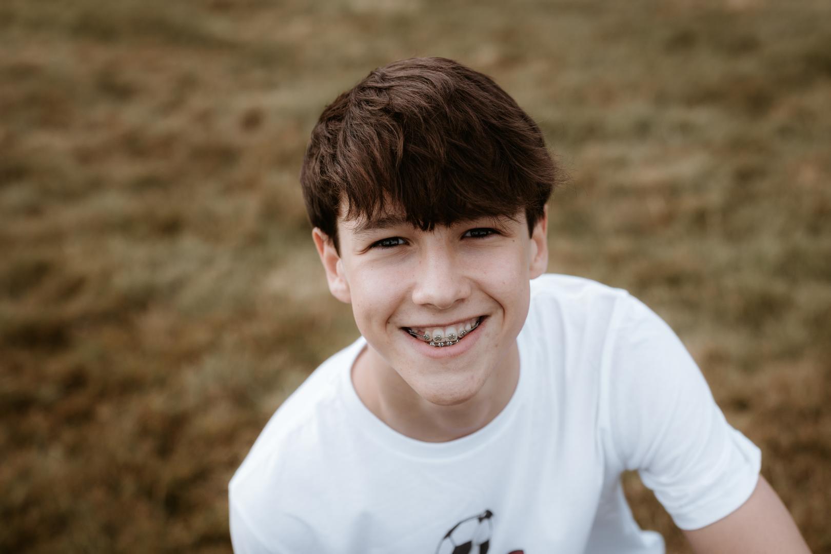 A teenage boy with braces smiles at the camera, with grass behind him