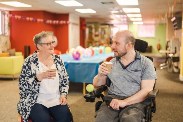 An older white woman with glasses sits in a community centre drinking tea and smiling to a younger white man in a wheelchair sitting next to her, who is also drinking tea.