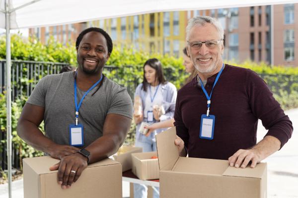 Freepik - a dark skinned young man in a grey shirt and a white skinned older man in a long black t-shirtare standing outside both holding boxes. They are smiling at the camera and are wearing lanyards that say Volunteer on them. 