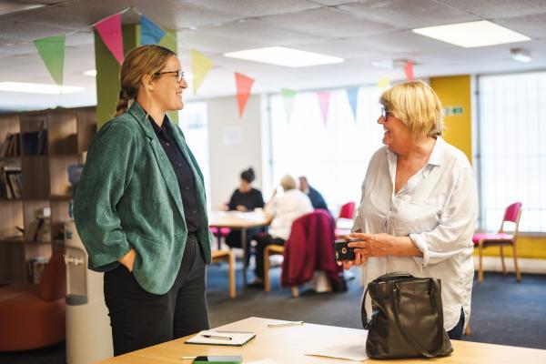 An image of the inside of a community centre showing two white women, one younger with long braided blonde hair, another older with short blond hair. They are standing speaking to each other in front of a table.