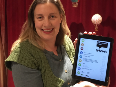 A smiling woman in a green jumper holds up an iPad with text that reads Carbon Literacy Award